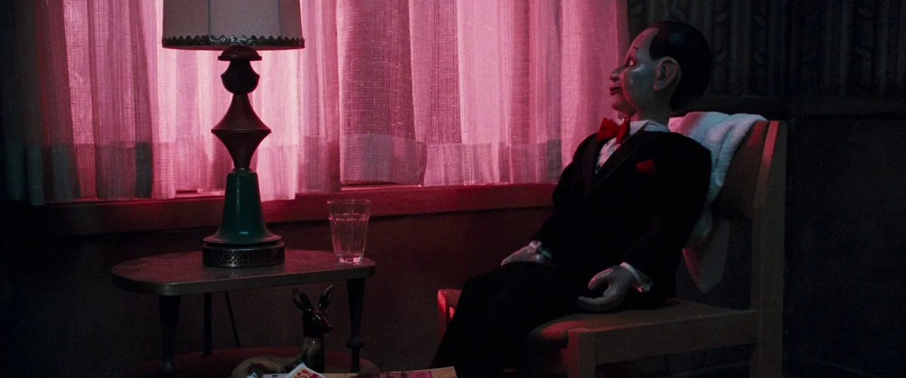 Dead Silence (2007) movie still: medium — A dark, dimly lit room has a ventriloquist dummy seated in a wooden chair. The background is dominated by…