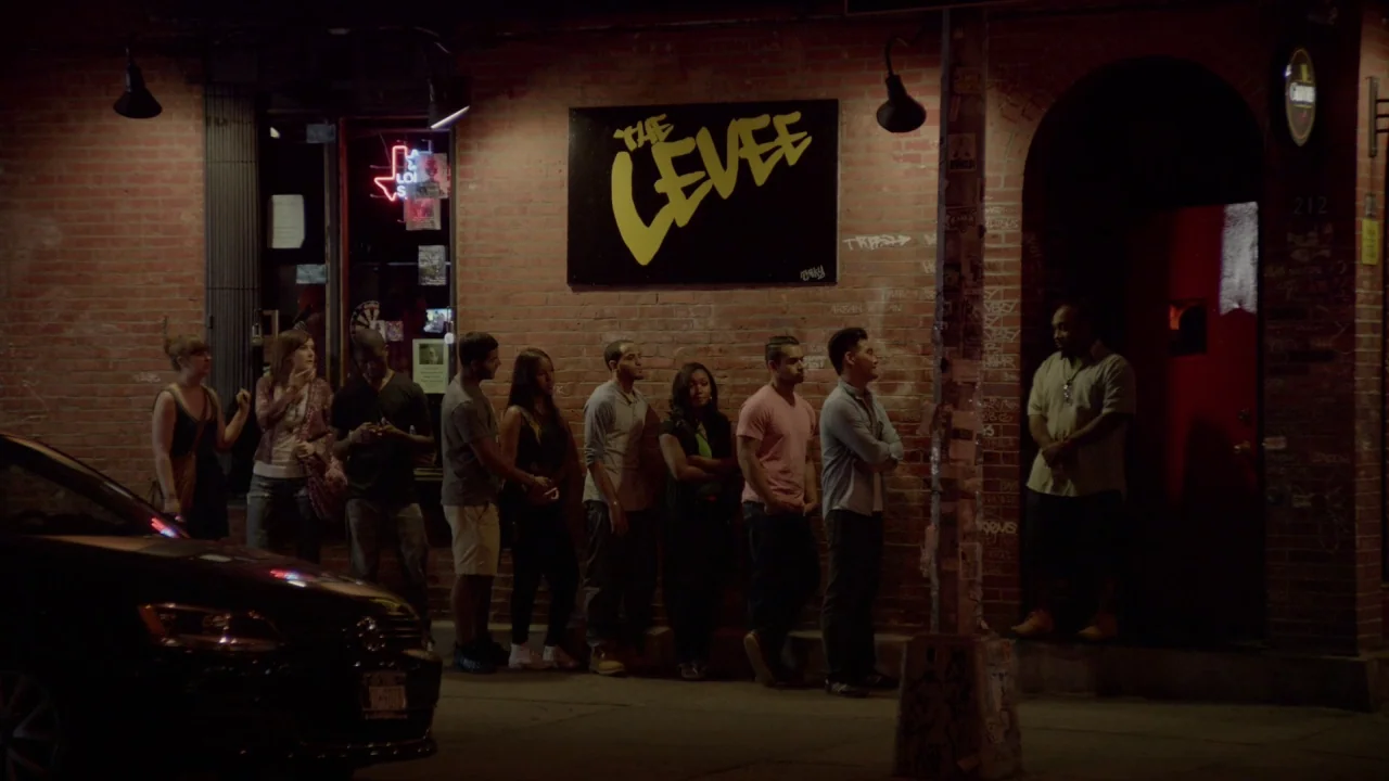 The Heart Machine (2014) movie still: wide — A brick-walled building exterior features a prominent yellow neon sign reading The Levee. A group of…
