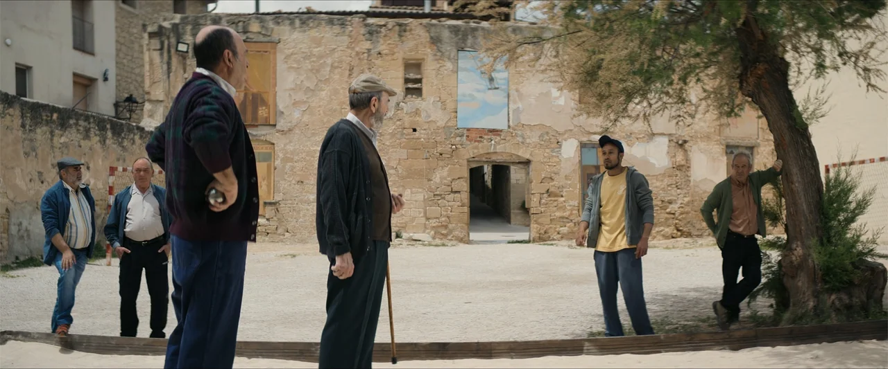 A Hipster in Rural Spain (2024) movie still: fullbody — A rural outdoor gravel court is surrounded by old stone walls and a tree. A group of men of…