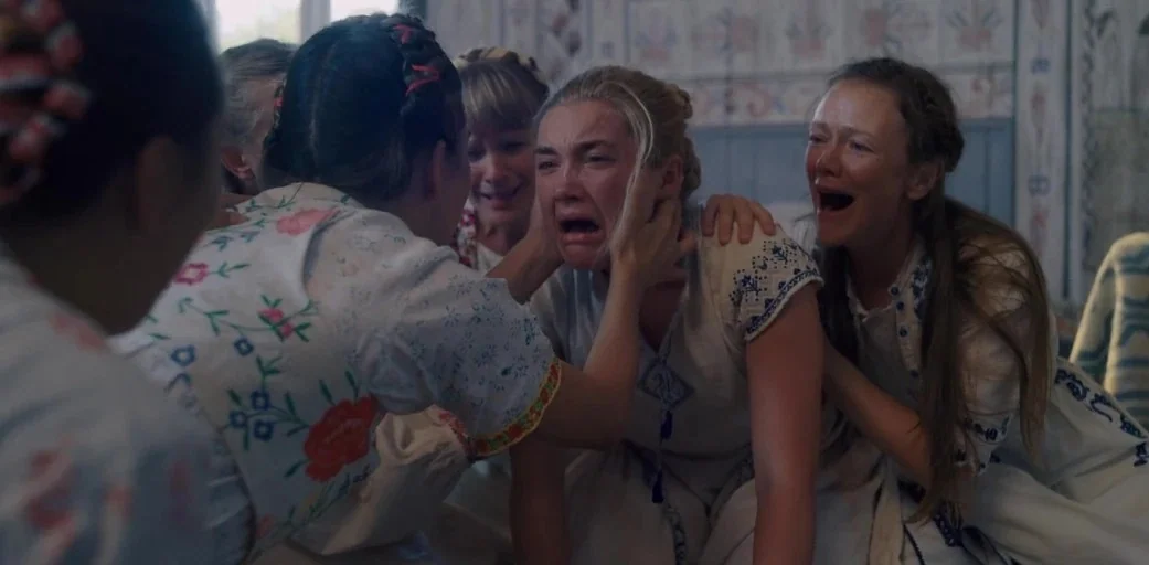 Midsommar (2019) movie still: medium — A group of women in embroidered white clothing gather in a light-colored interior room. The women hold onto a…