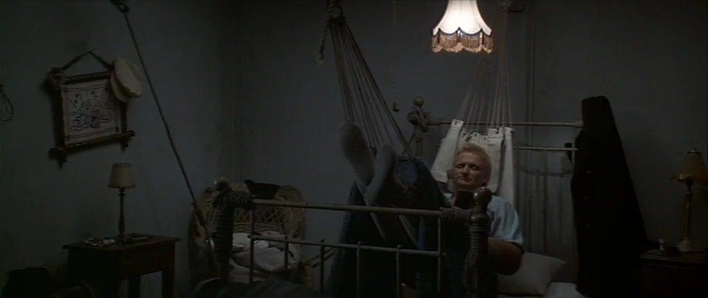 Popeye (1980) movie still: medium — A dimly lit room with walls painted in a cool, neutral tone. A man rests in a metal frame bed with his feet…