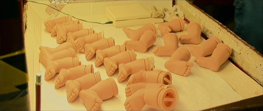 Bubble (2005) movie still: medium — A wooden work surface is covered with dozens of molded plastic infant doll legs. The prosthetic limbs are…