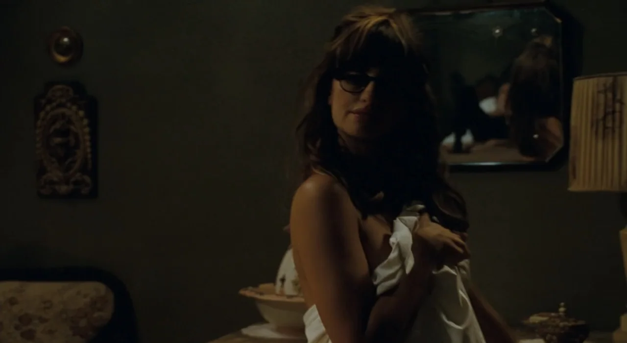 The Great Beauty (2013) movie still: medium — A dark bedroom features a wall-mounted mirror, a table lamp, and antique decor. A woman covered by a…