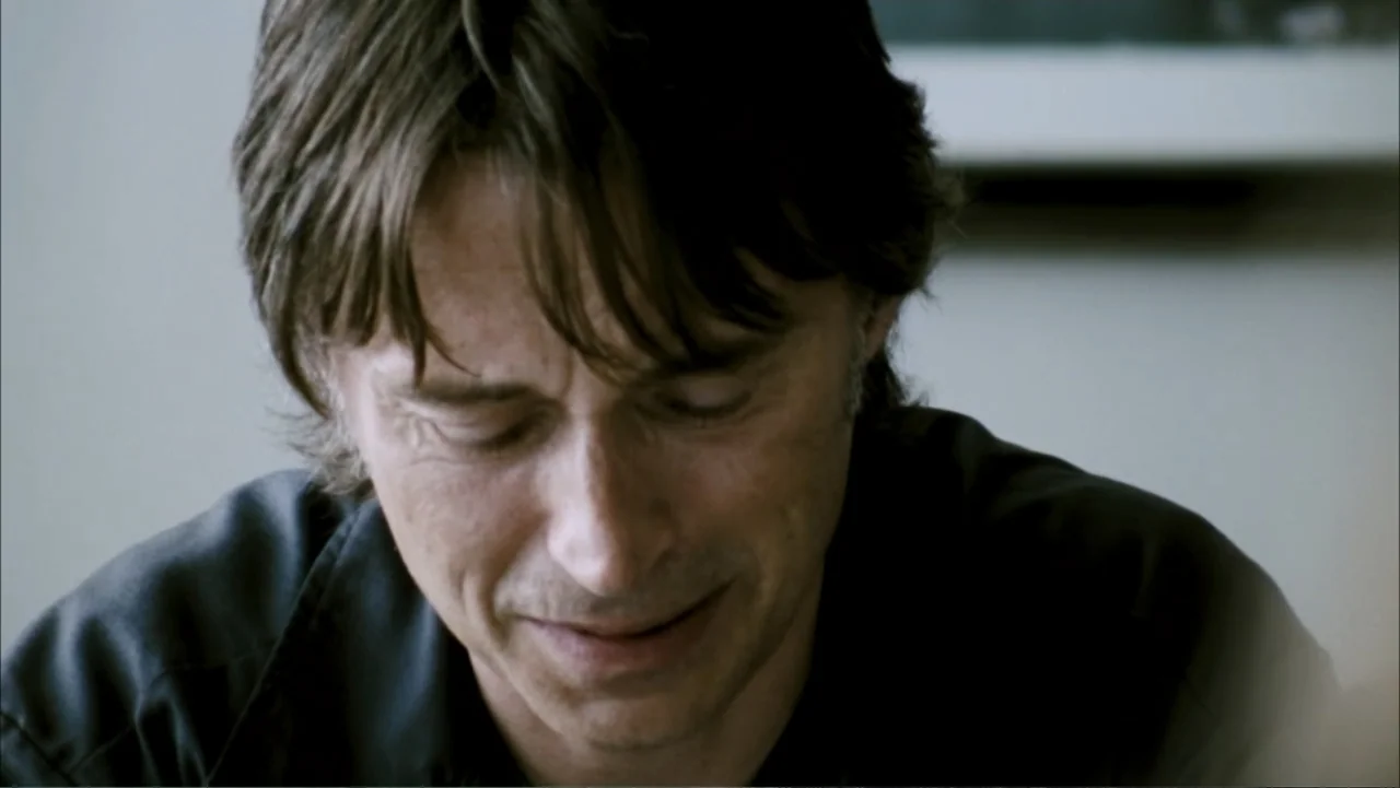 28 Weeks Later (2007) movie still: closeup — A man stands in a dimly lit room with plain walls. He has his eyes tightly closed and his facial…