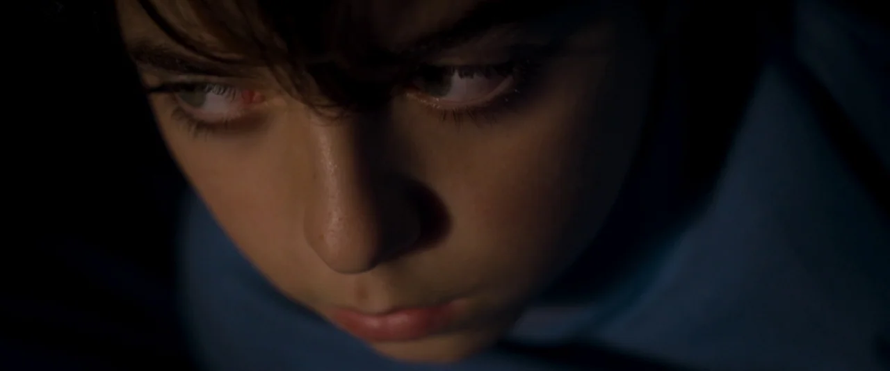 Baskin (2015) movie still: closeup — A dimly lit interior space is dominated by dark surroundings and near-black shadows. The face of a young person…
