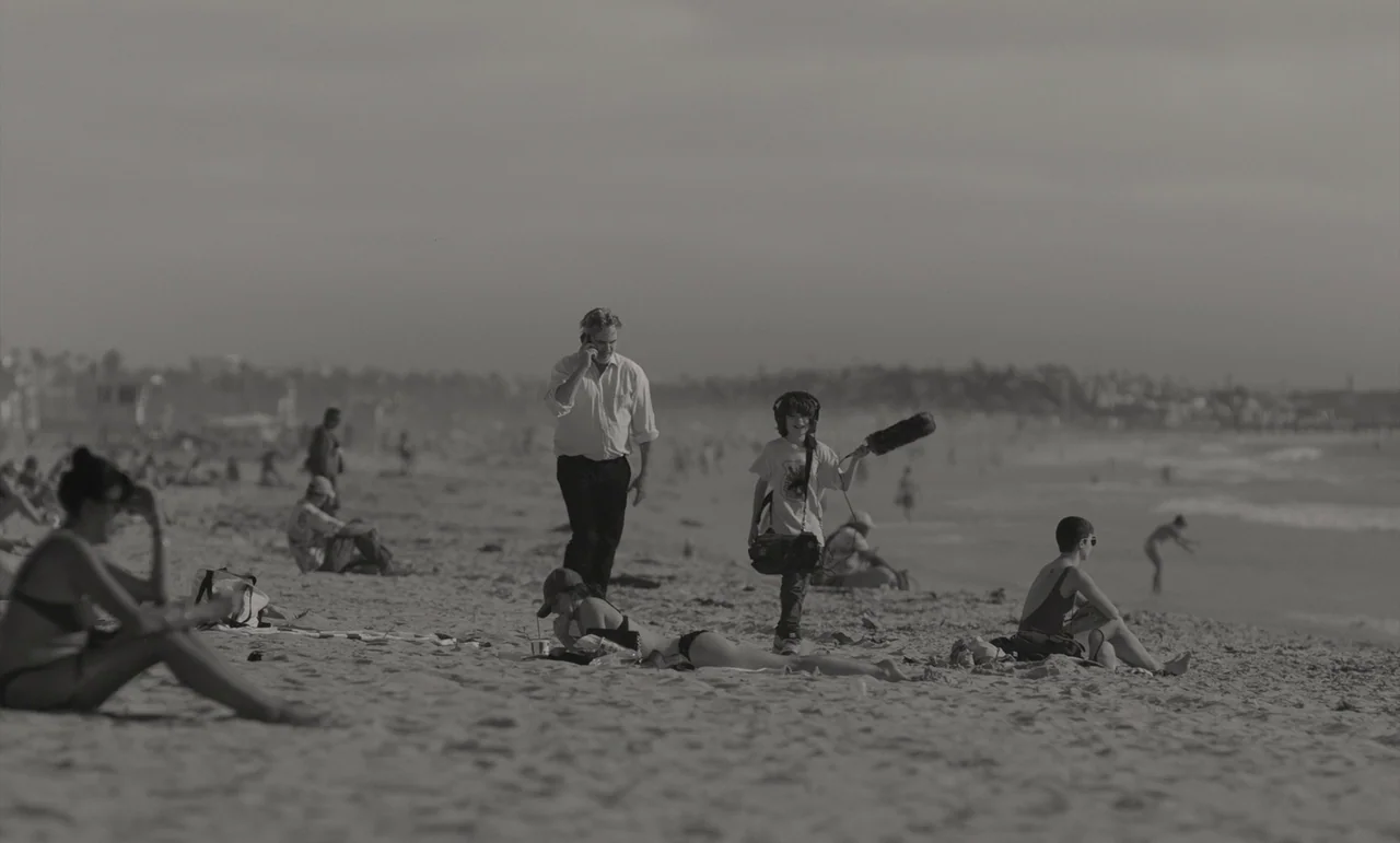 C’mon C’mon (2021) movie still: wide — A wide shot of a public sandy beach filled with people relaxing near the water. A man in a dress shirt talks…