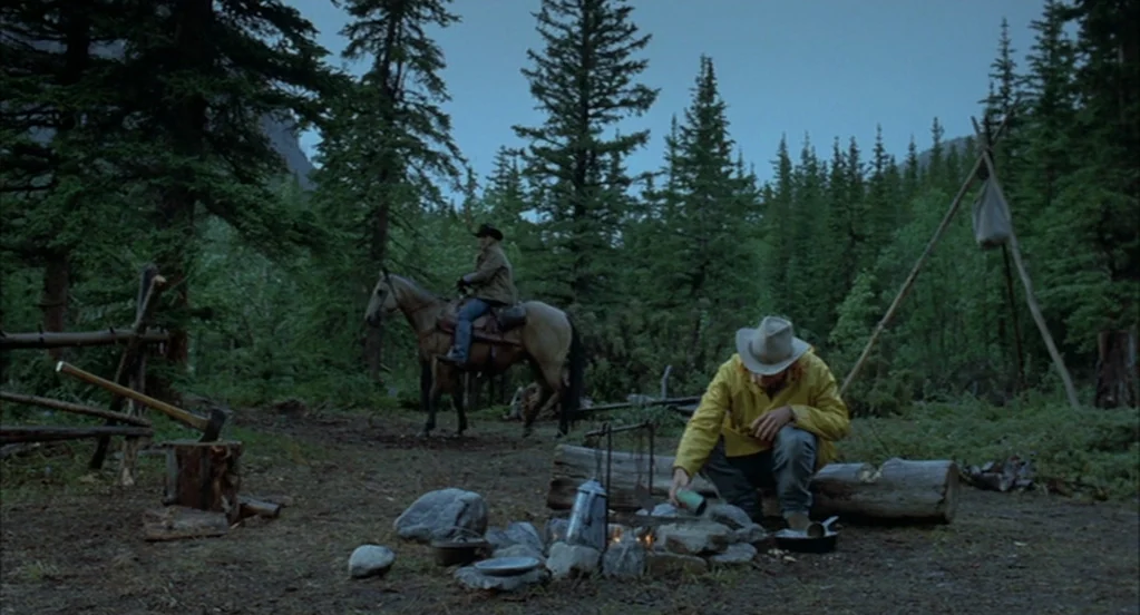 Brokeback Mountain (2005) movie still: wide — A remote campsite in a dense pine forest features a central stone fire pit. A man in a yellow jacket…