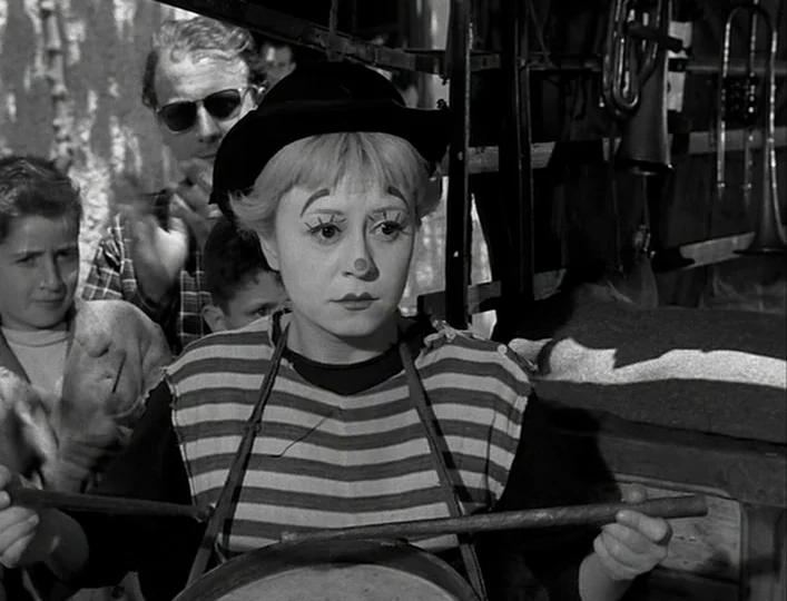 La Strada (1954) movie still: medium — A woman in clown makeup stands before a rack of musical instruments outdoors. She holds drumsticks over a…