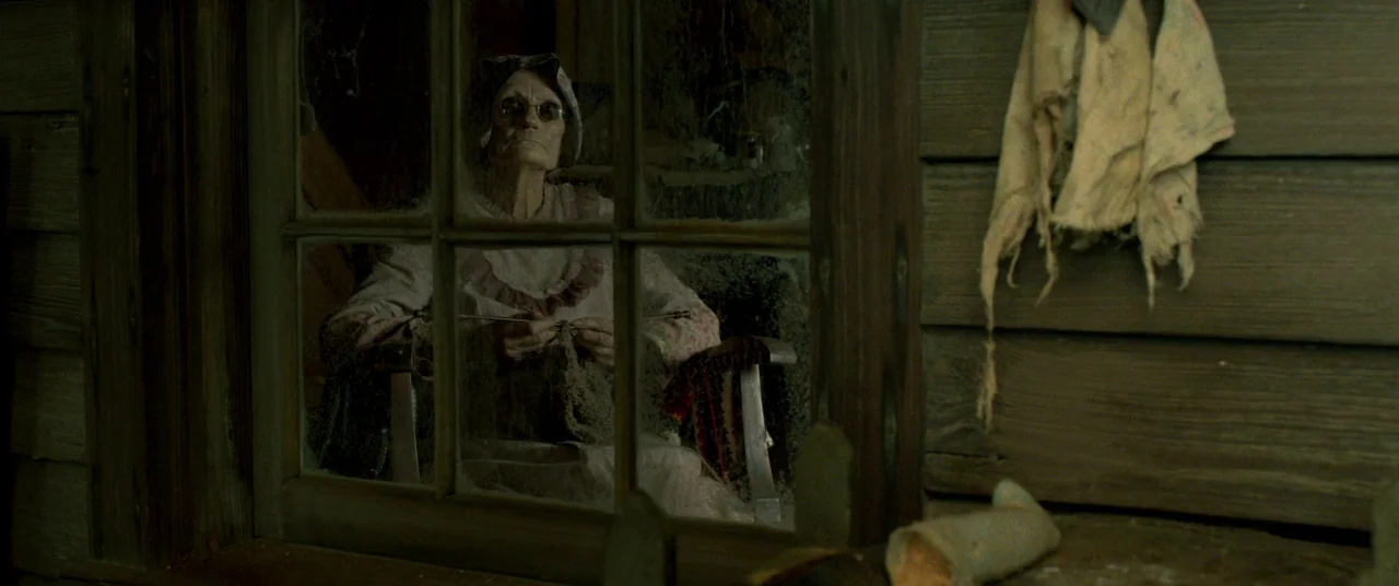 Lawless (2012) movie still: medium — An elderly woman sits behind a pane of glass in a dark wooden structure. She holds knitting needles while…