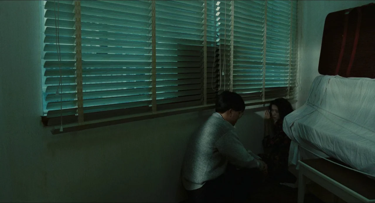 Rouge (1987) movie still: wide — A dimly lit, sparse room with horizontal window blinds and a white-covered object nearby. A man and a woman sit low…