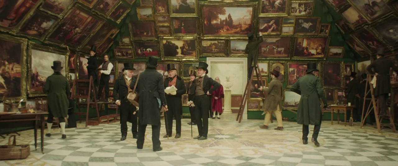 Mr. Turner (2014) movie still: wide — An 1800s art gallery has green walls densely packed with framed oil paintings. Men in period tailcoats and top…