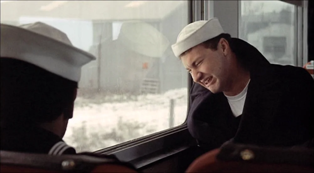 The Last Detail (1973) movie still: medium — Two sailors in uniform sit inside a passenger train car looking out a window. Outside the window, a…