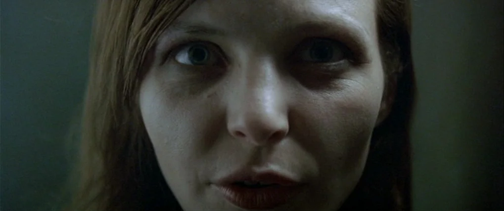 Next Door (2005) movie still: closeup — A face is illuminated in an otherwise dark, undefined interior space. The subject gazes directly forward…