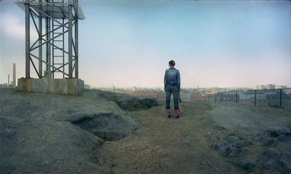 You, The Living (2007) movie still: fullbody — A rocky outcrop overlooks an expansive cityscape under a clear blue sky. A lone figure stands on the…