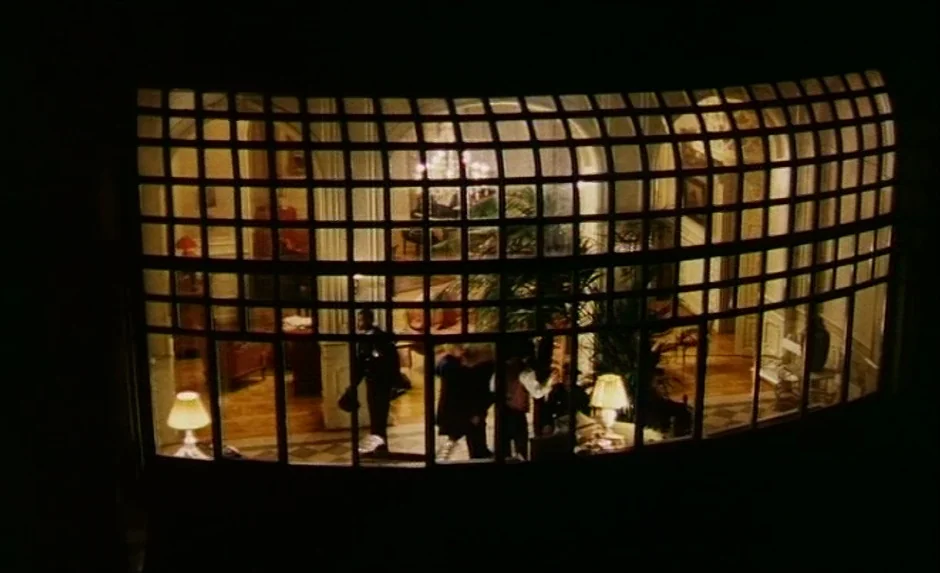 Simon, The Magician (1999) movie still: wide — A dark exterior view captures a large curved window wall divided by a grid. Silhouetted figures move…