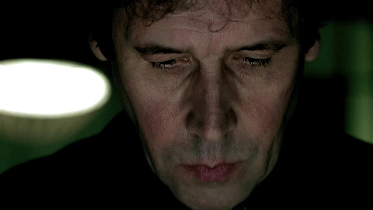 In Dreams (1999) movie still: closeup — A man's face is illuminated in a dark room with a blurred light source nearby. He looks down with his head…