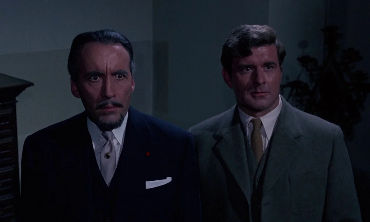 The Devil Rides Out (1968) movie still: medium — A dimly lit room with plain walls frames two men in formal attire. The subjects stand together…