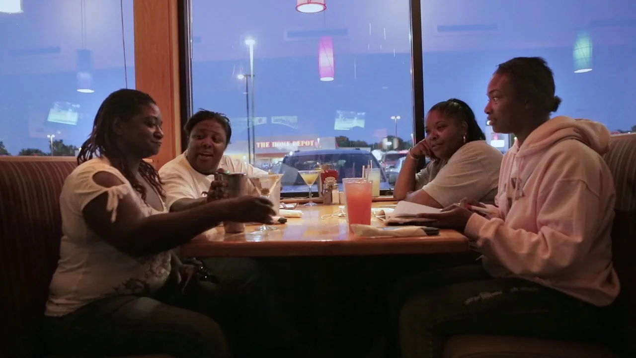 T-Rex (2015) movie still: medium — A restaurant booth with a wooden table holds glasses and drinks during the evening. Four women sit in the booth…