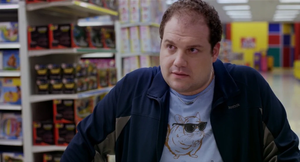 Dark Horse (2011) movie still: medium — A well-lit retail toy aisle features tall shelves stocked with various boxed products. A man in a graphic…
