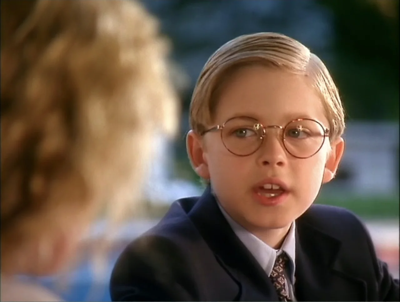 At the Midnight Hour (1995) movie still: closeup — A sunny park setting with soft-focus greenery in the background. A young boy in a blazer speaks…