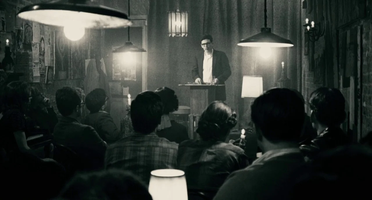 Howl (2010) movie still: medium — A dimly lit room functions as a gathering space with exposed brick and posters. A man stands at a wooden podium…