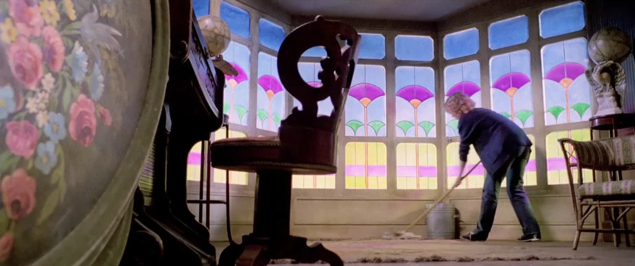 House By The Cemetery (1981) movie still: wide — A brightly lit sunroom features expansive stained glass windows with purple and green floral…