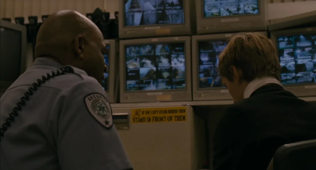 Restless (2011) movie still: medium — A dark, cramped surveillance room features stacks of monitors displaying multi-camera feeds. A security guard…