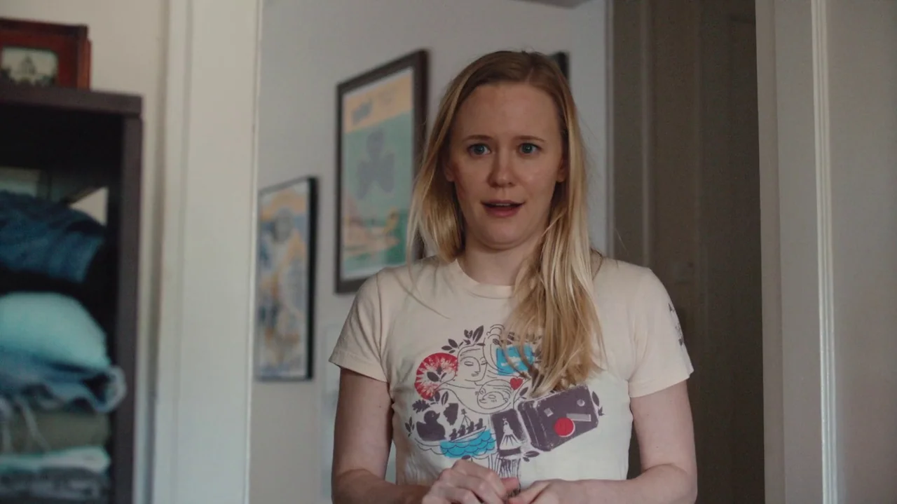 Saint Frances (2019) movie still: medium — An interior residential space features white walls adorned with framed art prints. A blonde woman stands…