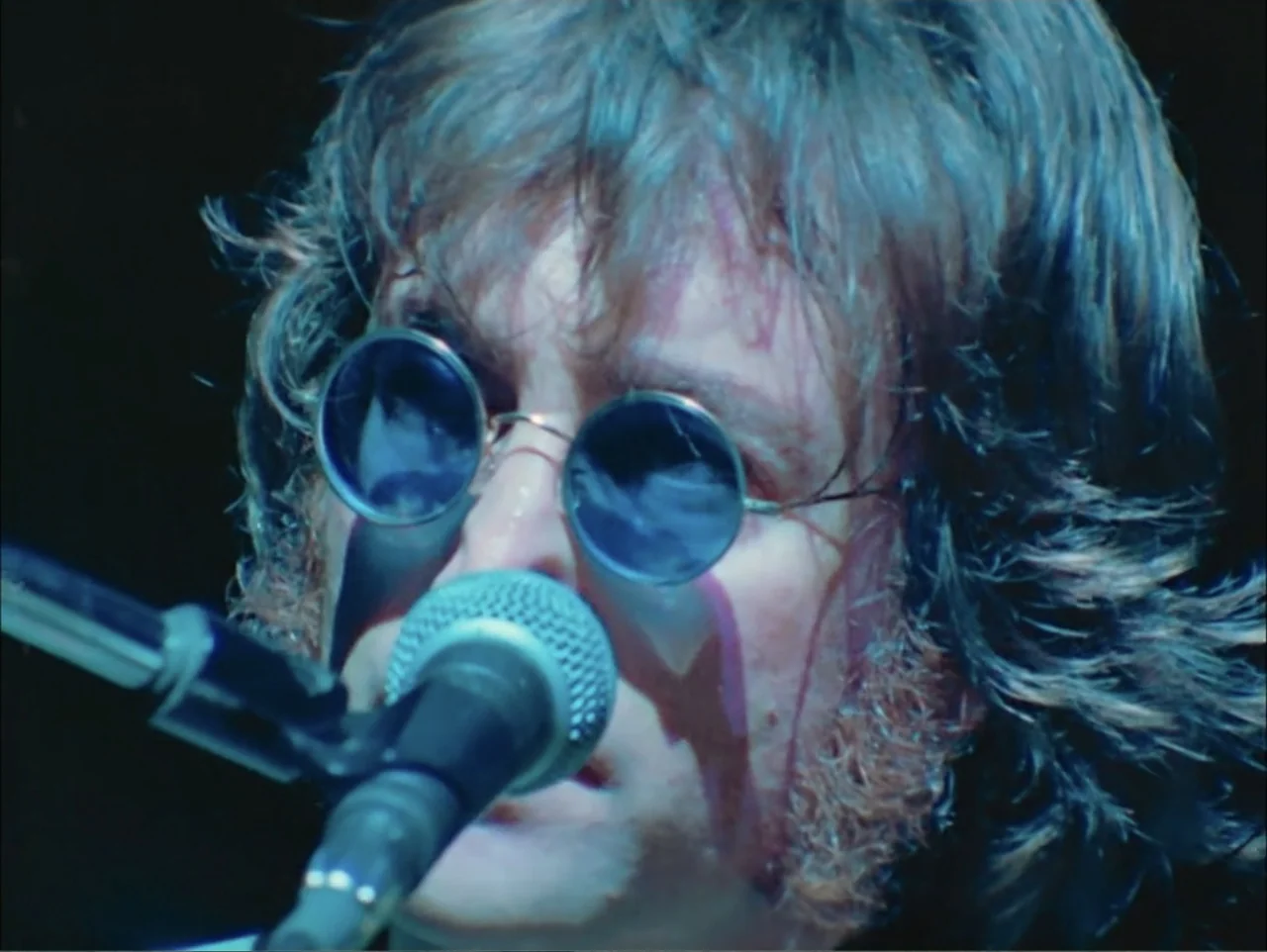 One to One: John & Yoko (2025) movie still: closeup — A dark recording studio interior with a black background. A man with shoulder-length hair and…