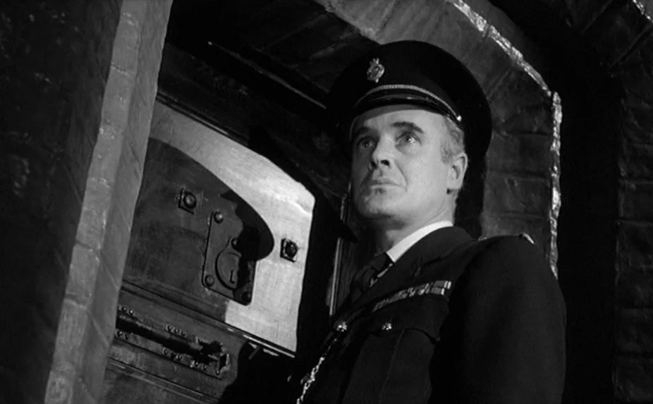 The Criminal (1960) movie still: medium — A stone corridor featuring a thick, recessed metal door with a small viewing slot. A uniformed official in…