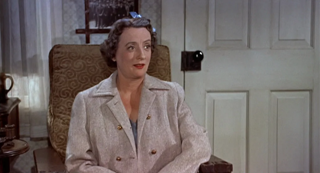 The Trouble With Harry (1955) movie still: medium — A classic living room interior features an armchair set against a white door. The woman sits in…