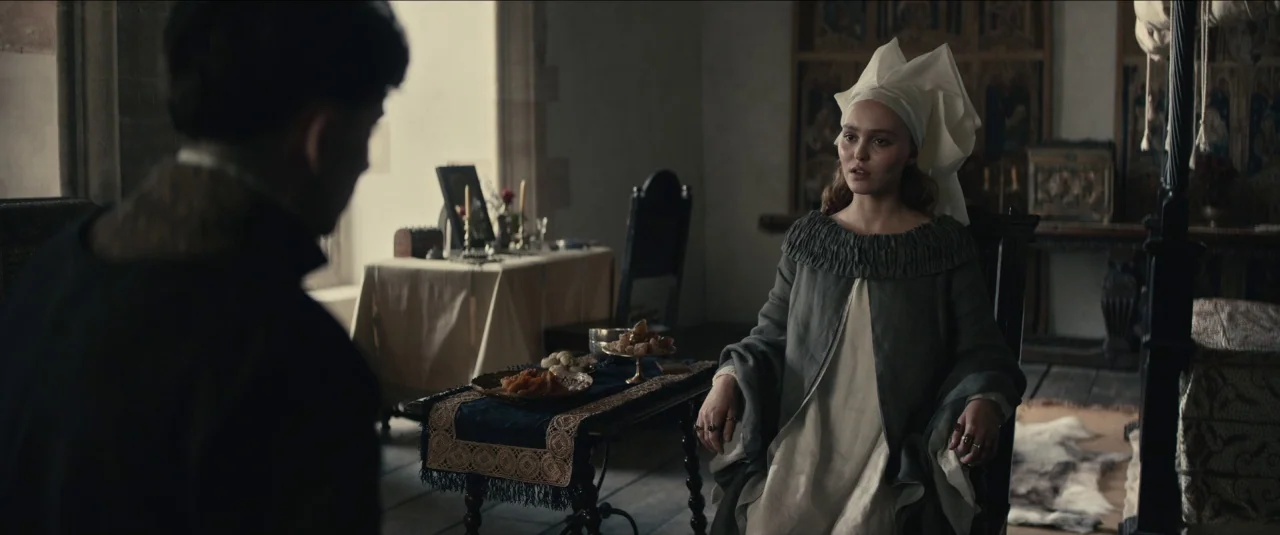 The King movie still: medium — A stone-walled medieval room features a wooden table with fruit and a draped bed. A young woman in a white headdress…