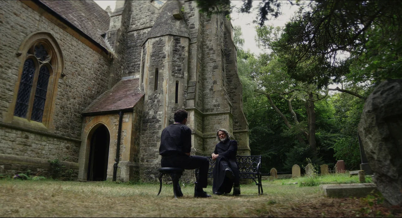 Wake Up Dead Man (2025) movie still: wide — A stone church building stands adjacent to a grass field with scattered tombstones. An elderly man and a…