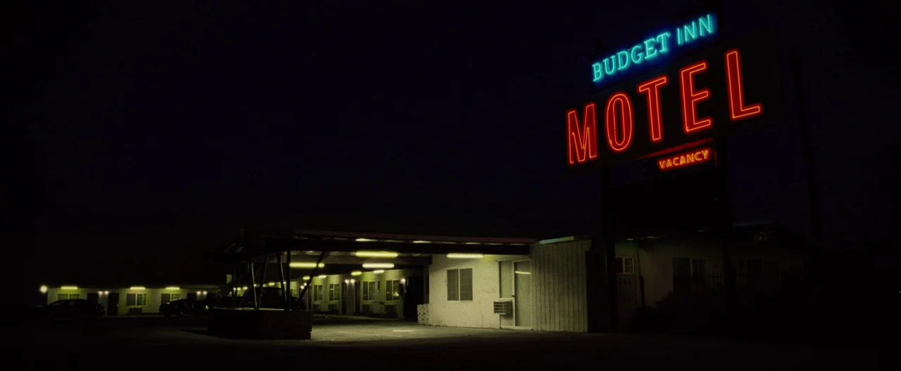 Nocturnal Animals (2016) movie still: wide — A dark night exterior features a glowing neon sign for a Budget Inn motel. The building has a carport…