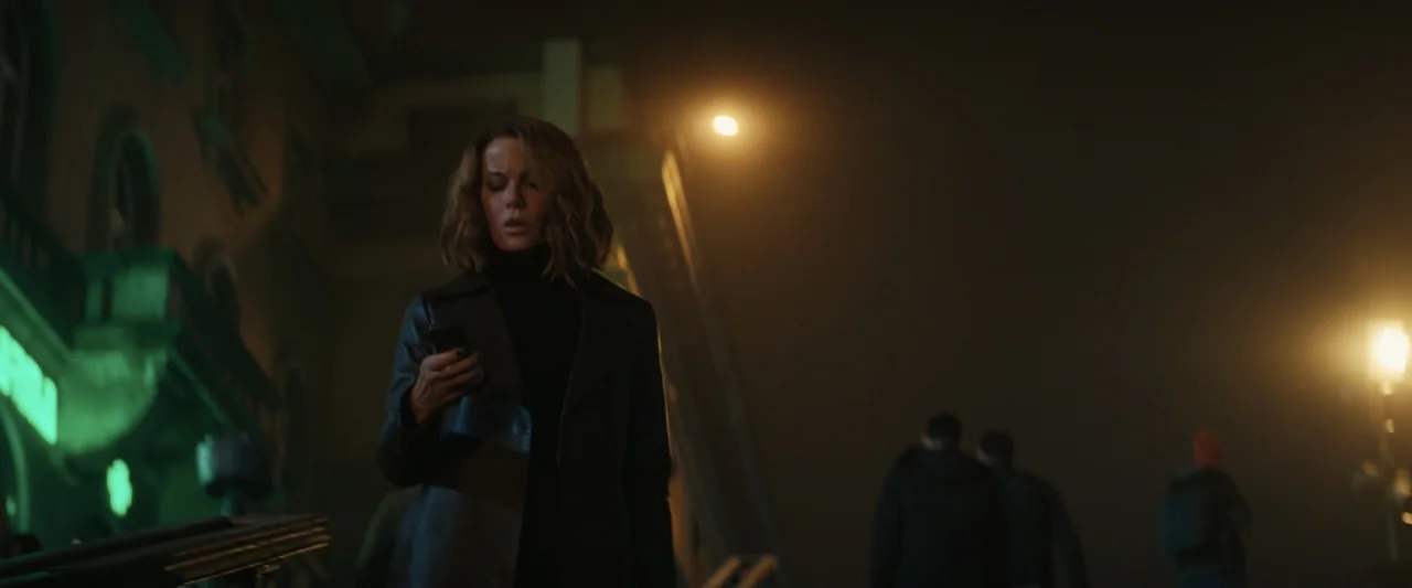Canary Black (2024) movie still: medium — A dark urban street at night with glowing streetlights and green building illumination. A woman standing…