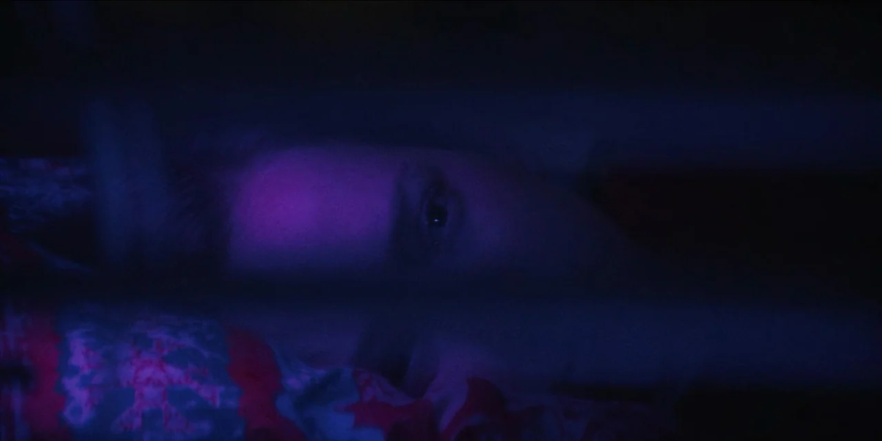 Spermworld (2024) movie still: closeup — A dark bedroom interior is bathed in shifting blue and magenta light. A person lies in bed with only one…