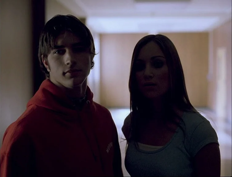 Elephant (2003) movie still: medium — A dimly lit hallway with a bright window at the far end. A young man and woman stand in the foreground against…