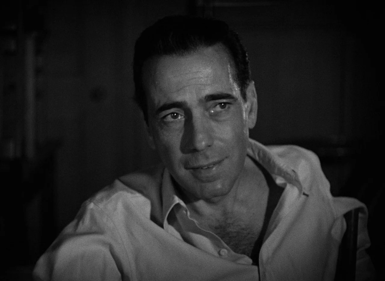 Key Largo (1948) movie still: closeup — A man is centered in a dimly lit room with a dark background. He sits wearing an unbuttoned white shirt…