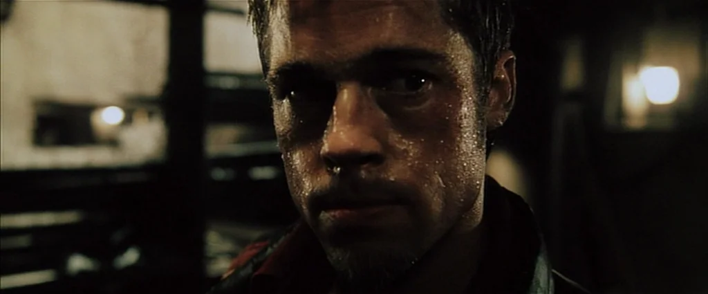 Fight Club (1999) movie still: closeup — A dark, dimly lit space with deep shadows and visible industrial features. A man with a sweaty face stares…