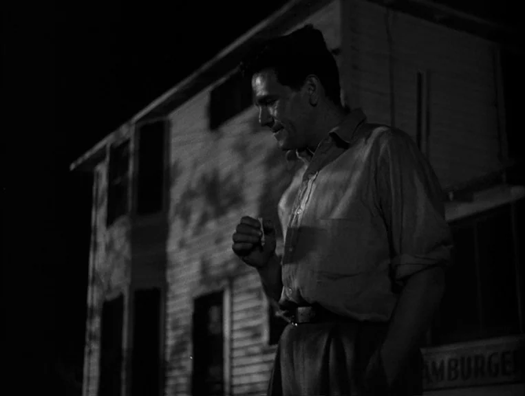 The Postman Always Rings Twice (1946) movie still: medium — A dark night scene outside a wood-sided building with faint text visible. A man stands…