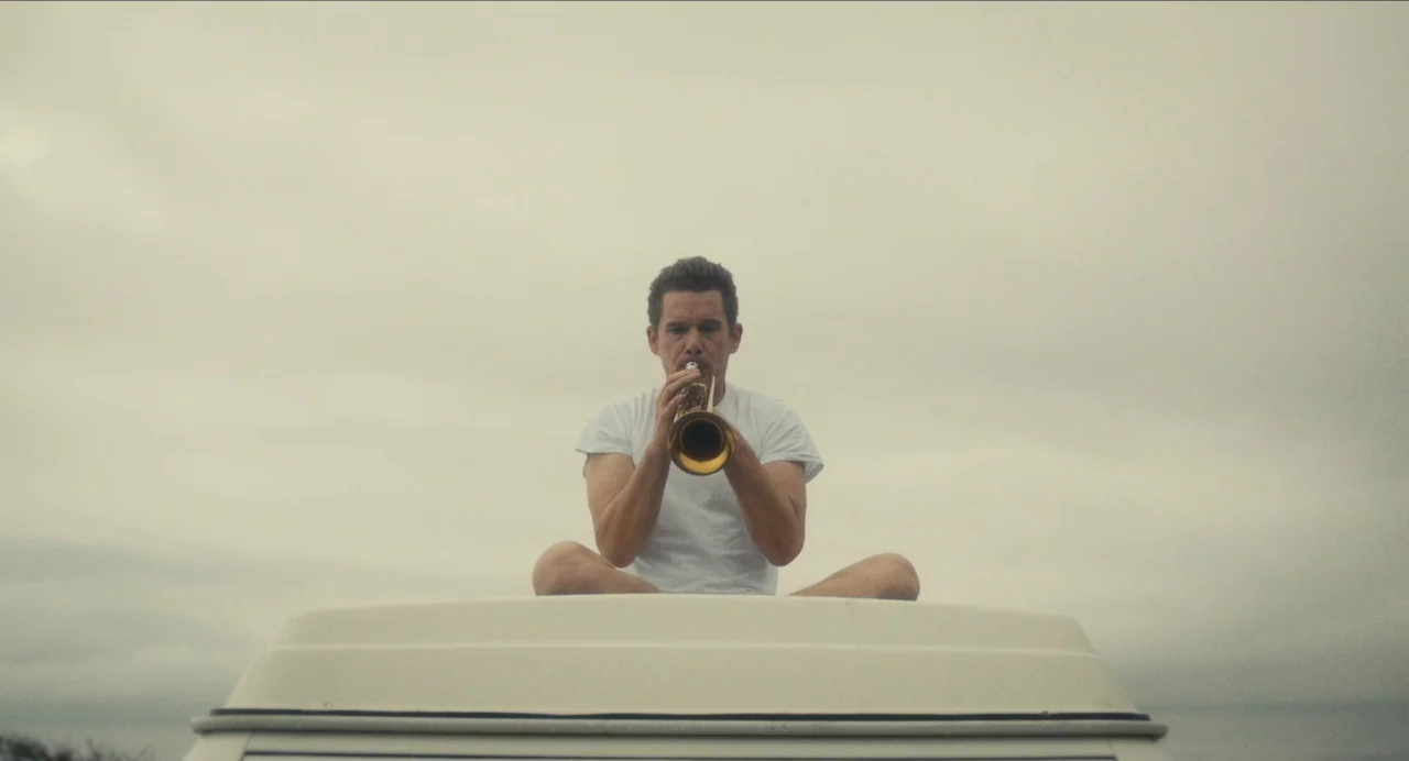 Born To Be Blue (2015) movie still: medium — A man sits cross-legged on the flat white roof of a vehicle outdoors. The backdrop is a vast, overcast…
