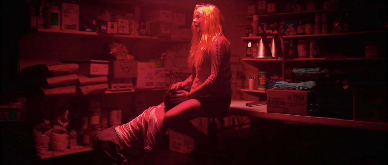 68 Kill (2017) movie still: medium — A cluttered storage room filled with shelves of various boxes and products. A young woman sits on a table as a…