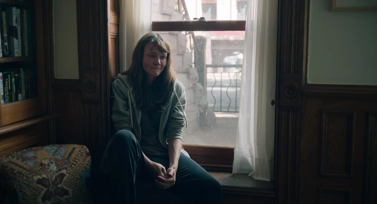 She Said (2022) movie still: medium — A wooden window seat is situated between a bookcase and a window looking outdoors. A young woman sits on the…