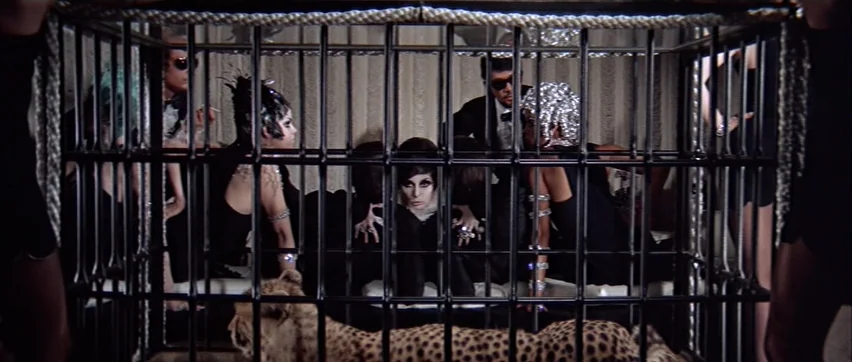Sweet Charity (1969) movie still: medium — A group of people sits inside a large metal cage in a furnished room. A cheetah lies on the floor at the…