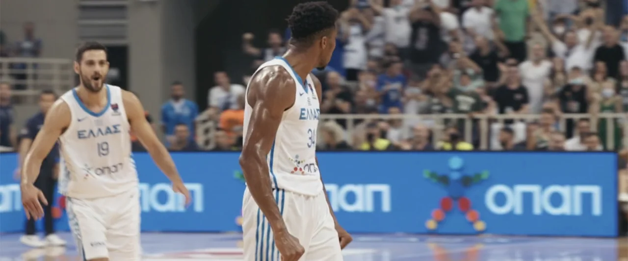 Giannis: The Marvelous Journey (2024) movie still: medium — A brightly lit indoor sports arena with a blue advertising board in background. Two…