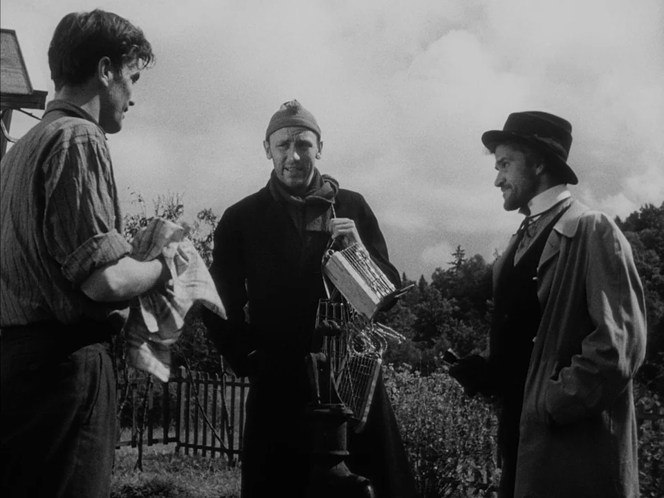 It Rains On Our Love (1946) movie still: medium — A rural setting with a wooden fence and distant trees under an overcast sky. Three men gather near…