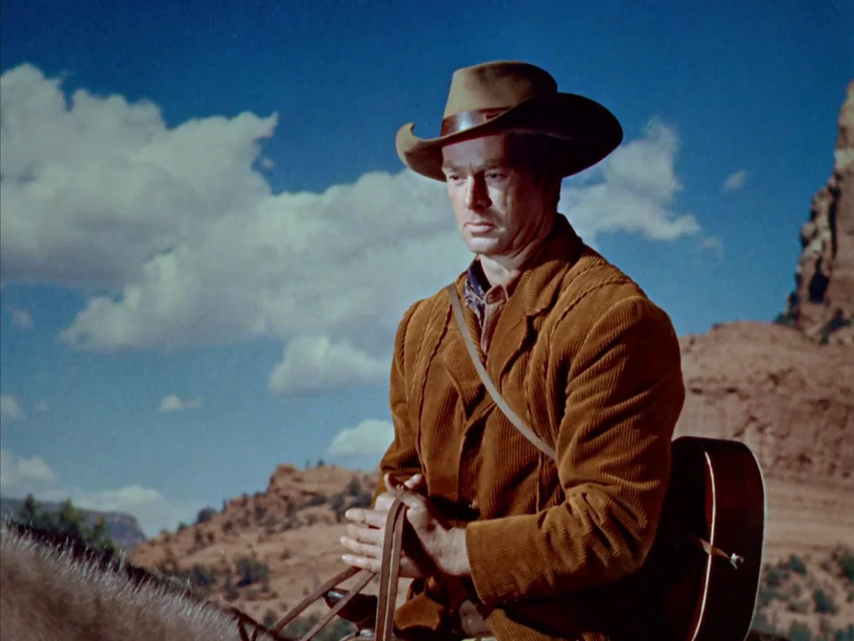 Johnny Guitar (1954) movie still: medium — A rugged desert landscape features red rock formations under a bright blue sky. A man on horseback holds…