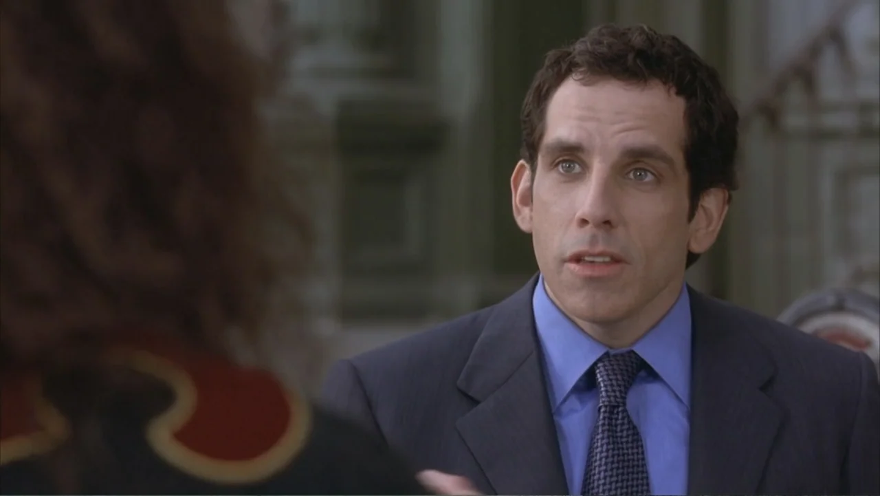 Along Came Polly (2004) movie still: medium — A man in a suit stands in a diffuse daylight setting. The man engages in conversation while viewed…