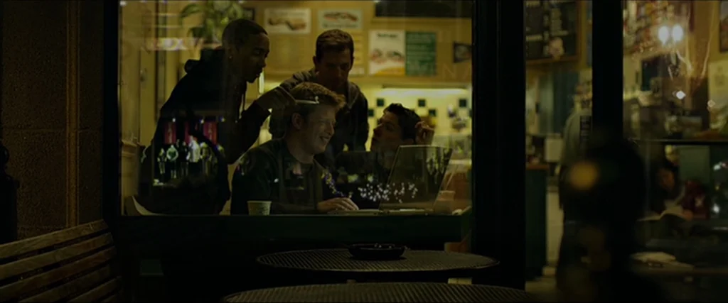 The Social Network movie still: medium — Three young men cluster around an open laptop on a table inside a dimly lit venue. The view is partially…