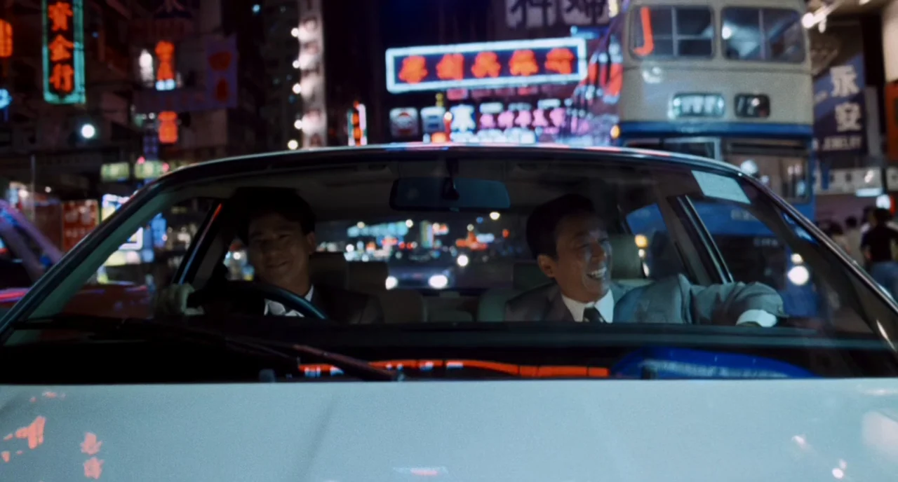 The Killer (1989) movie still: medium — A night view from inside a car driving through a neon-lit Hong Kong street. Two men in business attire sit…